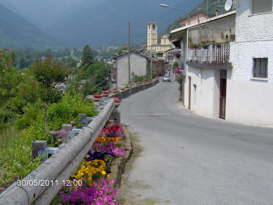 Villar Pellice (TO). La via centrale è costeggiata a sinistra da aiuole fiorite. Come sfondo le montagne ricoperte di vegetazione verde 
