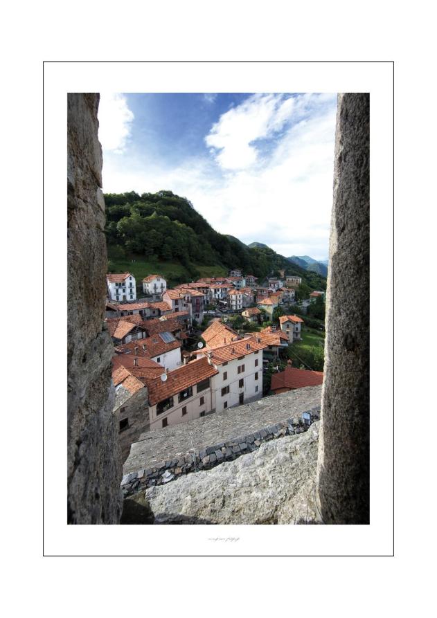 Arola (VCO). Da una finestra in pietra si vedono i tetti del paese e le montagne in lontananza. Il cielo è nuvoloso. Fotografia di Enzo Franza (2013) 