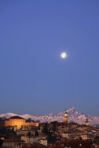 Saluzzo (CN). In lontananza si vede il paese illuminato dalle prime luci serali. Il cielo è limpido e la luna brilla in alto. Sullo sfondo le montagne innevate. 