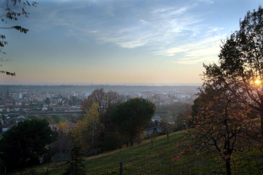 Vigliano Biellese (BI). Panoramica mentre albeggia. Il sole è ancora basso e nascosto fra gli alberi. Il paese si intravede in lontananza. Immagine tratta dal sito del Comune 