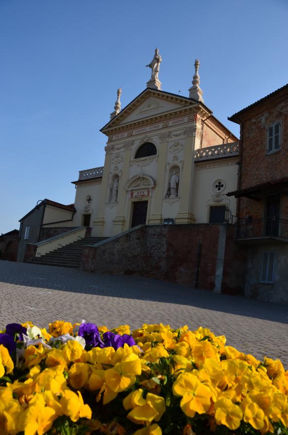 Scurzolengo (AT). In primo piano una aiuola di fiori gialli. In quadrata dal basso la Chiesa dei Santi Andrea e Lorenzo svetta nel cielo limpido e azzurro 