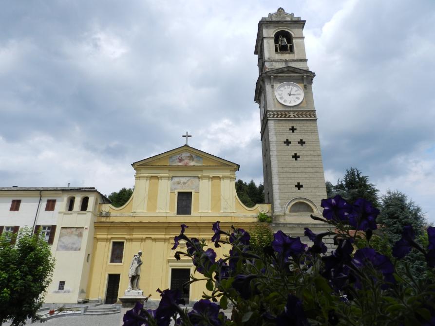 Bricherasio (TO). La facciata barocca della chiesa parrocchiale di Santa Maria Assunta è dipinta color crema . Alla sua destra il campanile grigio con orologio. Il cielo sullo sfondo è coperto di nuvole grigie 