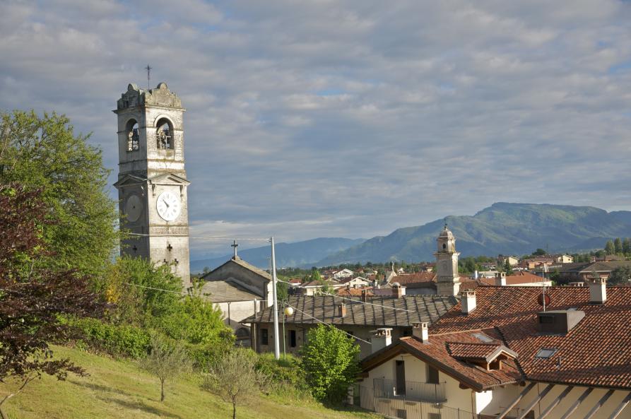 Bricherasio (TO). A sinistra svetta un campanile grigio fra la vegetazione. A destra spiccano i tetti del paese e in lontananza si intravedono i profili delle montagne. Il cielo è coperto di nuvole grigie 