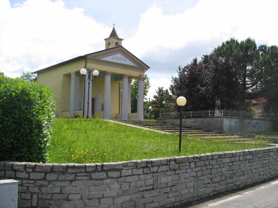 Caresana (VC). Un muretto di pietre grigie contiene un prato in salita che conduce alla Chiesa di Santa Maria. Quattro colonne bianche conducono all'entrata della chiesa. A destra un gruppo di alberi. Il cielo è nuvoloso. Fotografia di Claudio Tambornino (2008) 