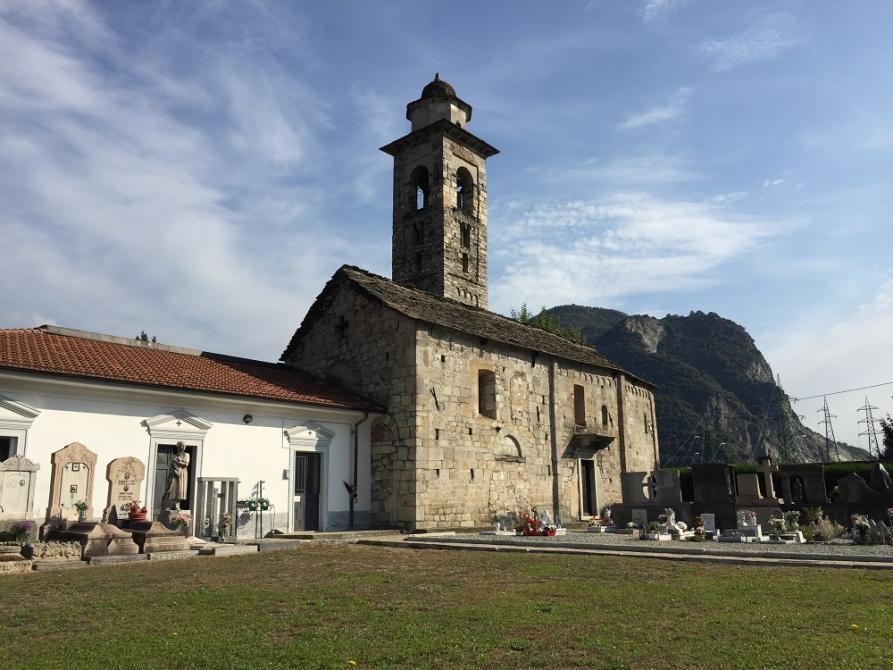 Gravellona Toce (VCO). La piccola Chiesa di San Maurizio, rivestita di pietre grigie, è fotografata di lato. Si intravedono lapidi funerarie  del cimitero limitrofo. Il cielo è azzurro e solcato da leggere nuvole bianche 
