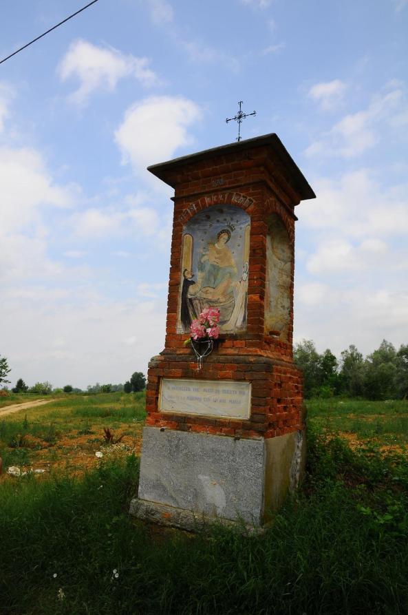 Sanfrè (CN). Lungo il sentiero verdeggiante compare un capitello religioso in mattoni scoperti con immagine sacra affrescata. Il cielo alle spalle è solcato da impalpabili nuvole bianche 