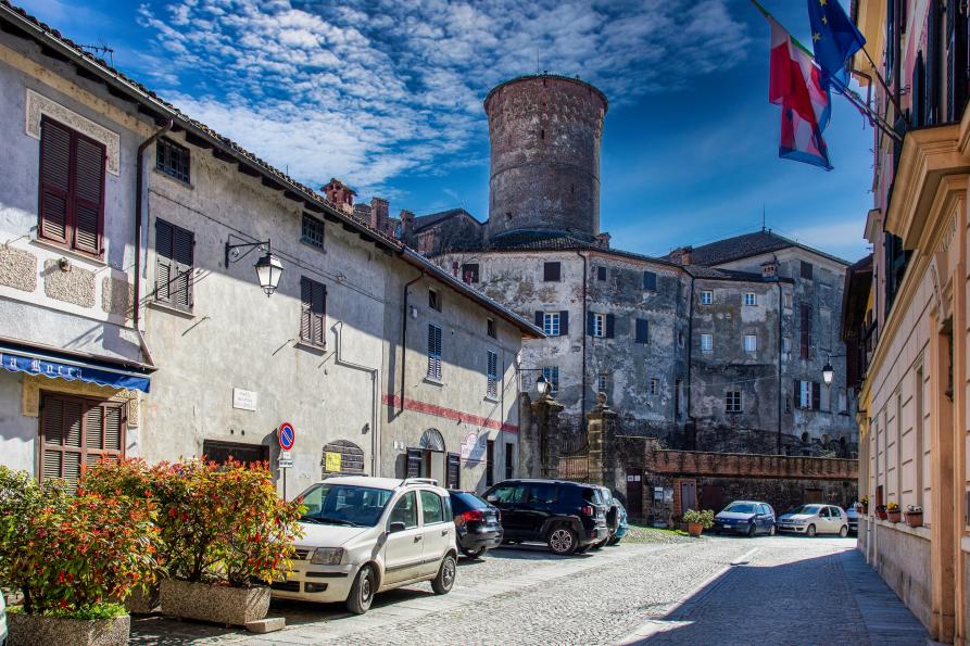 Rocca Grimalda (AL). Scorcio di Piazza Borgatta con auto parcheggiate sul lato sinistro e a destra i muri del Municipio con le bandiere. Al fondo si intravede il castello con la torre 