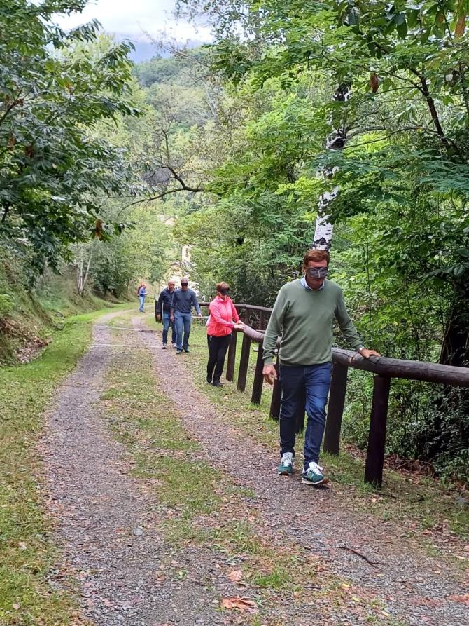 Vidracco (TO). Alcune persone camminano lungo un sentiero immerso nella natura con gli occhi coperti da una mascherina aiutandosi appoggiando le mani sulle recinzioni in legno 