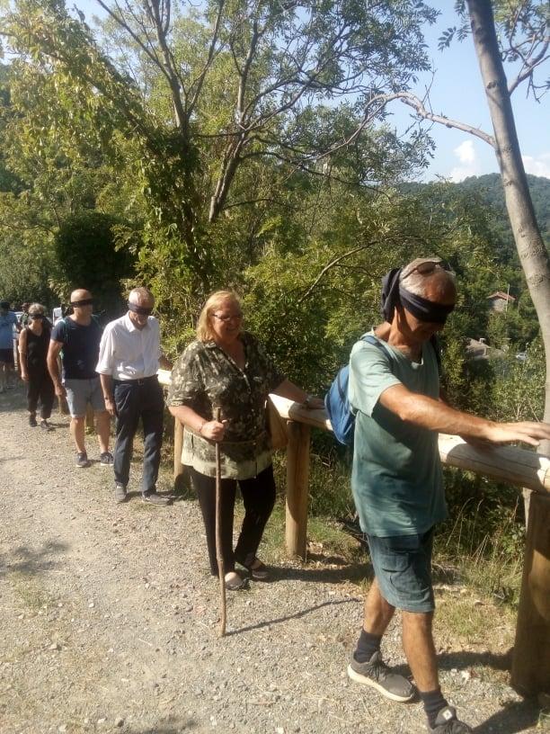 Vidracco (TO). Alcune persone sfilano lungo un sentiero sterrato. Hanno gli occhi coperti con una mascherina e camminano aiutandosi appoggiando le mani sulle staccionate di legno 