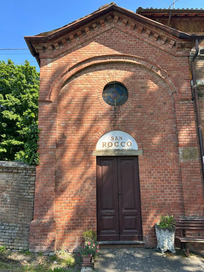 Castelletto Molina (ST). In primo piano la facciata in mattoni rossi con portale in legno sormontato da piccolo rosone in vetro della chiesetta di San Rocco 