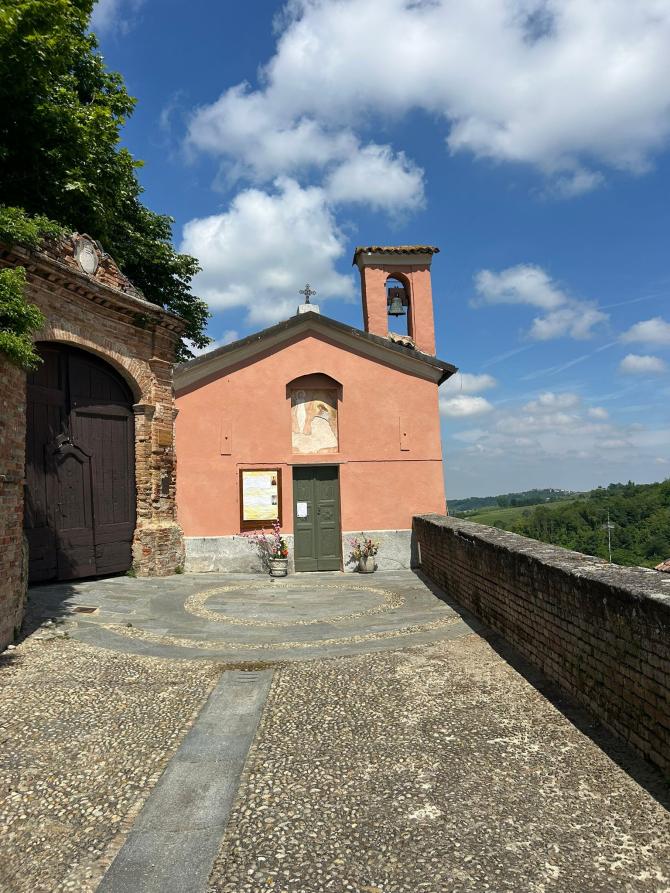 Castelletto Molina (AT). Lo spiazzo davanti all'Oratorio dell'Annunziata è lastricato con ciottoli. La facciata della chiesetta, dipinta color mattone, ha un piccolo portone di entrata in legno. La torre campanaria è a destra. Alle spalle il cielo blu nuvoloso 