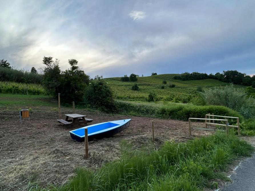 Castelletto Molina (AT). Tappa del percorso "Come una barca nelle vigne". Una barchetta azzurra accanto ad una panchina in legno. Intorno campi e colline verdi. Il cielo è nuvoloso  