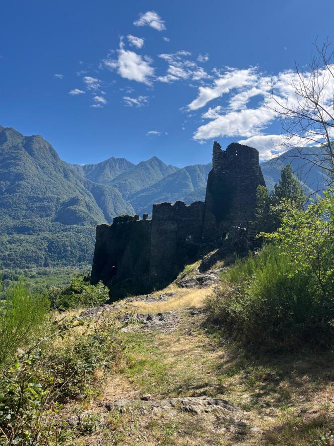 Vogogna (VCO). Il terreno della montagna è brullo e conduce a un rudere di un castello visto in silouette. Al fondo le alte montagne e il cielo blu con qualche nuvola bianca 