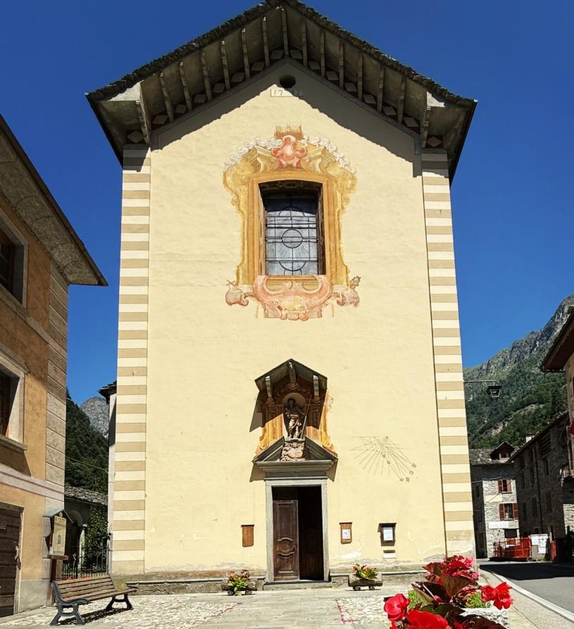 Mollia (VC). Facciata della Chiesa Parrocchiale dei Santi Giovanni e Battista con portone di ingresso in legno sovrastato da finestra rettangolare decorata. Il cielo alle spalle della chiesa è blu 