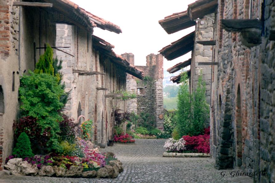 Candelo (BI). Ai lati della stradina in ciottolato le case in pietra viva a due piani del Ricetto sono abbellite e adornate di fiori . Al fondo si intravede l'interno di una torre merlata 