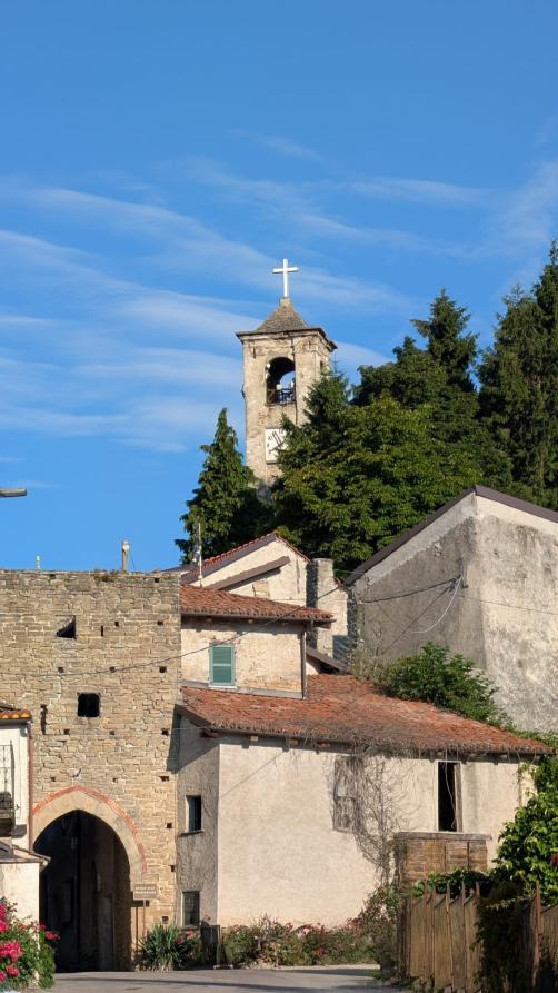 Mombarcaro (CN). In basso a sinistra l'Arco medievale in pietra a vista e alcune case addossate. Dietro alle cime degli alberi, a destra, spunta la torre campanaria. Il cielo è blu 