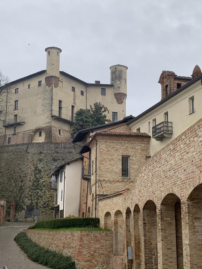 Cortanze (AT). In fondo a sinistra il castello si eleva rispetto alle case ritratte in basso a destra lungo le quali si dipanano arcate in mattoni rossi a vista 