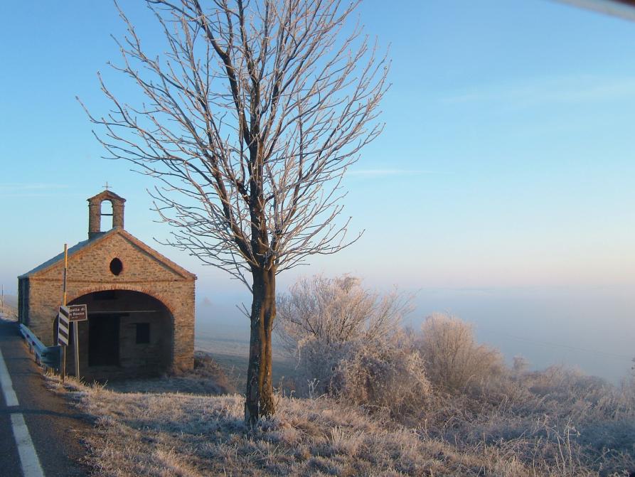 Mombarcaro (CN). A sinistra la facciata della chiesa di San Rocco in pietre e mattoni con ampio arco di entrata e piccolo campanile. Un albero spoglio sulla destra della chiesa e i prati e la vegetazione coperti di brina invernale. Il cielo è azzurro 
