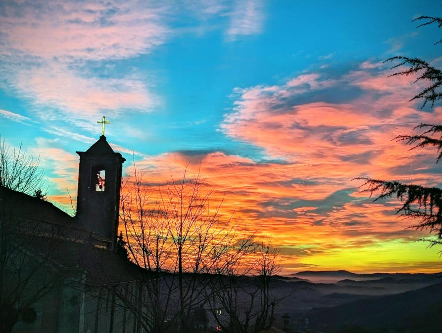 Mombarcaro (CN). A sinistra la figura in silhouette della Chiesa parrocchiale al tramonto. Il cielo azzurro ha nuvole tinte di arancione, rosa e giallo 