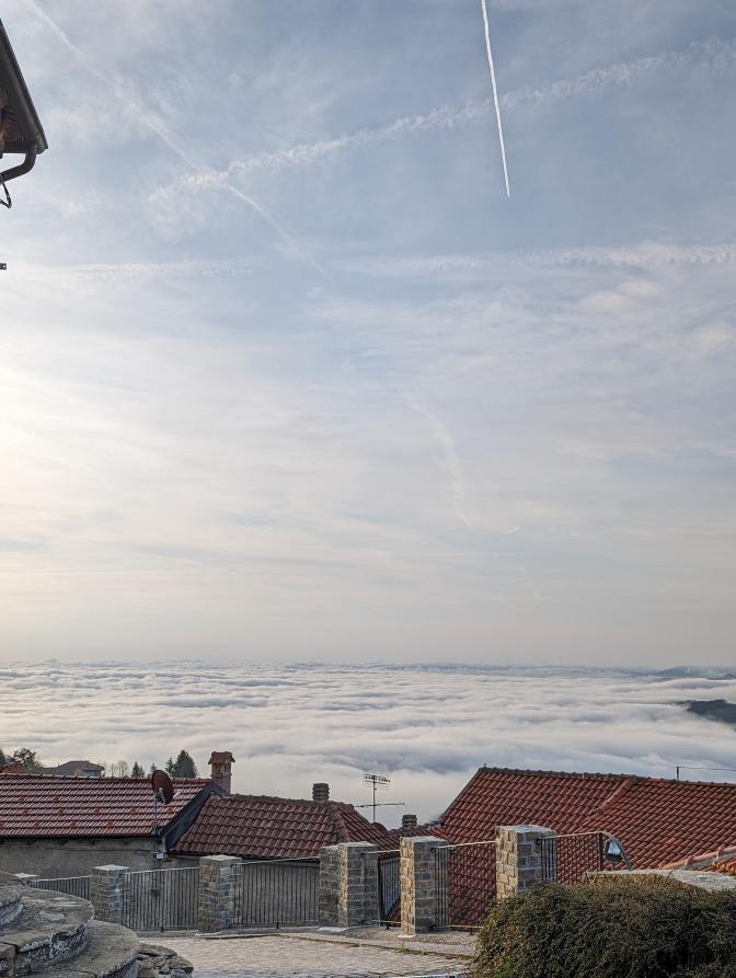 Mombarcaro (CN). Veduta da un terrazzamento su alcuni tetti del paese. La vista si perde sul mare di nebbia che si confonde con le nuvole grigie del cielo 